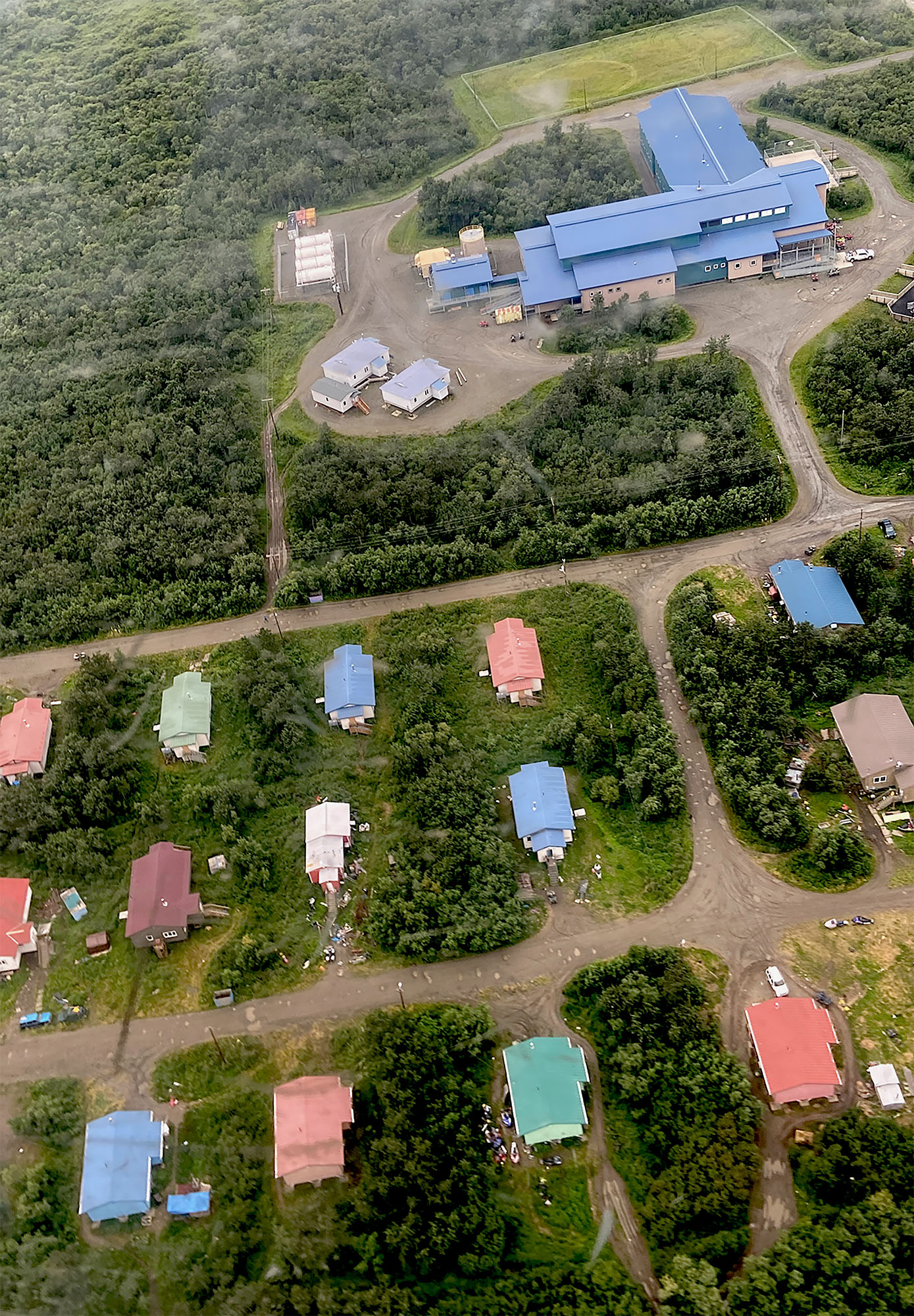 From an aerial view, surrounded by trees is a building with smaller structures in front of it.