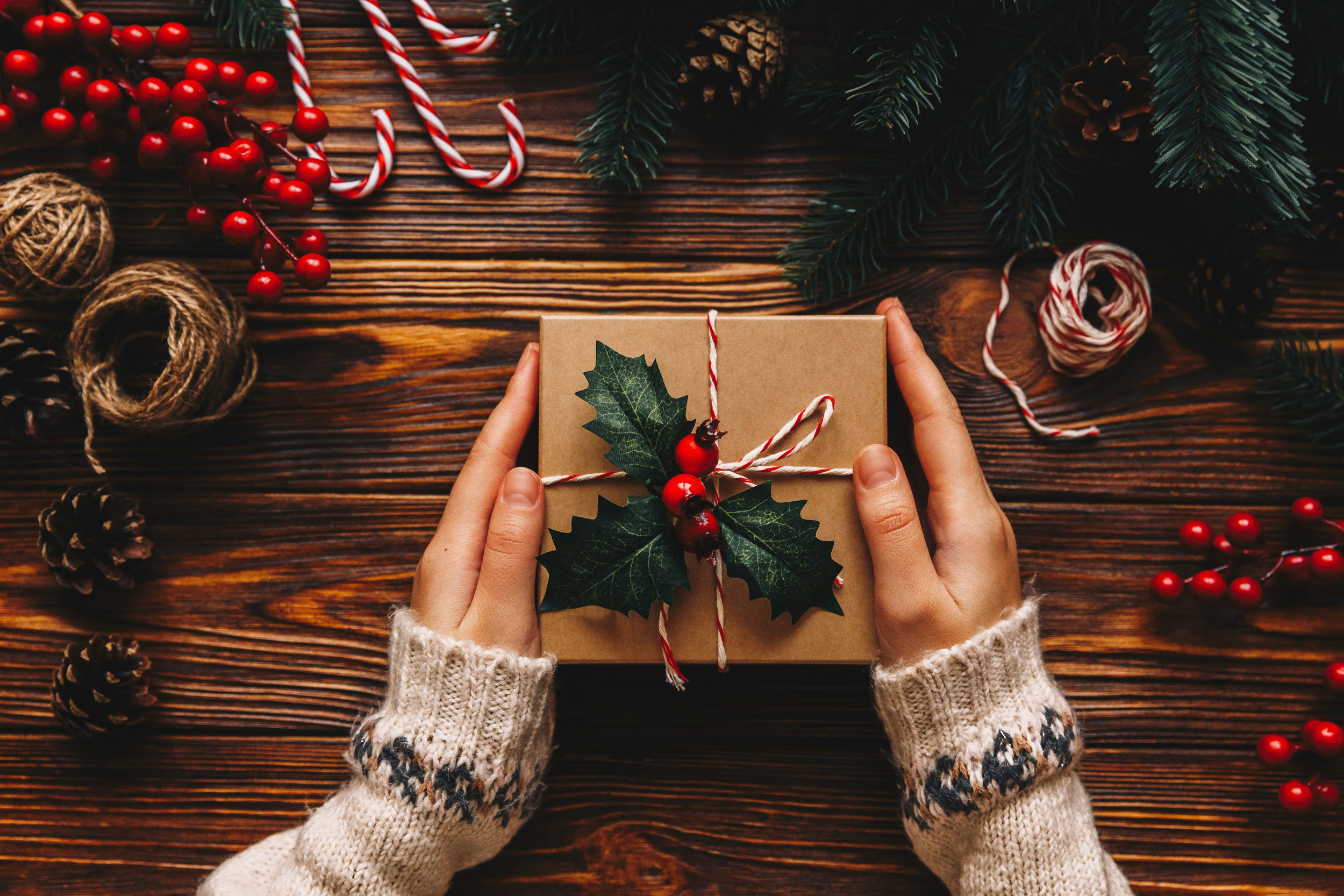 Adobe Stock image by Pink Coffee studio depicts a person holding a holiday package wrapped with holly. Other decorations are neatly stacked around the package, including pine branches, cones, twine, berries and sugar canes