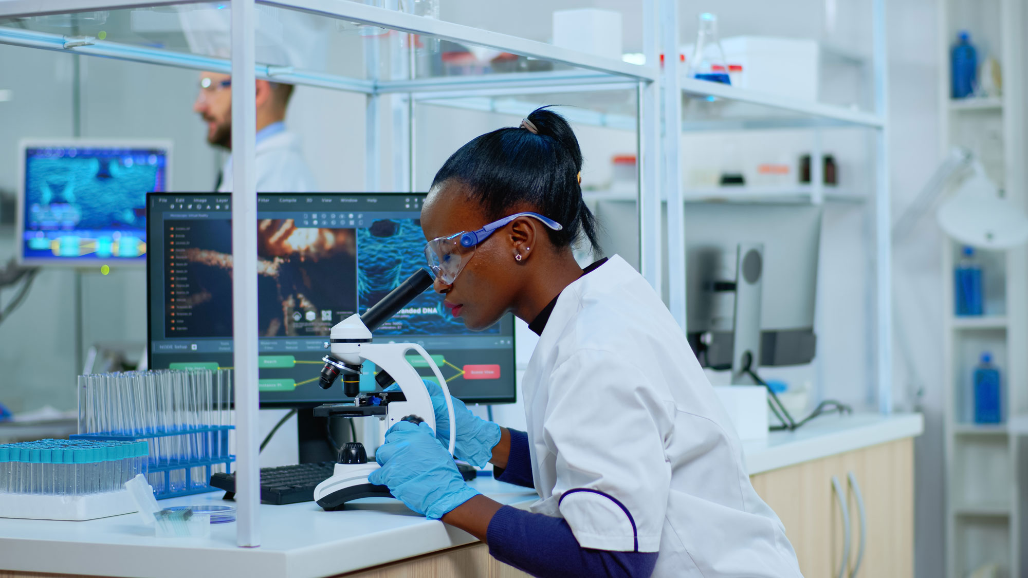 A scientist looks through a microscope in a laboratory. The slide she is observing is magnified on the monitor beside her