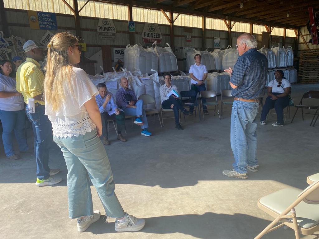 People in a delegation from Argentina, Paraguay, and Uruguay stand in a barn and sit front of bags of cover crop seed while listening to Bobby Hutchison discuss conservation efforts on his 130-year-old family farm
