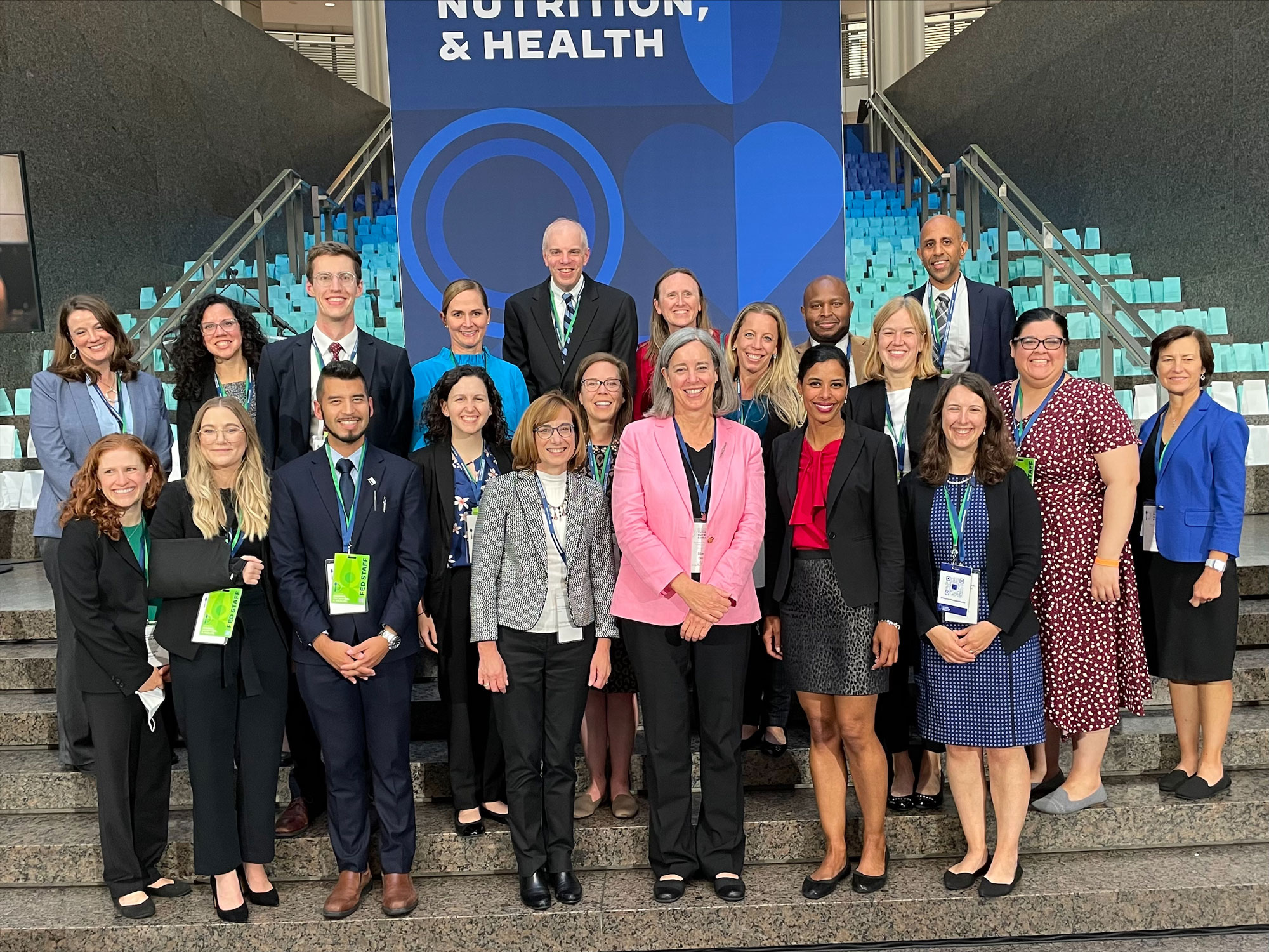 A group of 21 USDA Food and Nutrition Service employees dressed in professional attire and wearing conference badges pose together on a marble staircase in front of a blue conference banner at the White House Conference on Hunger, Nutrition, and Health