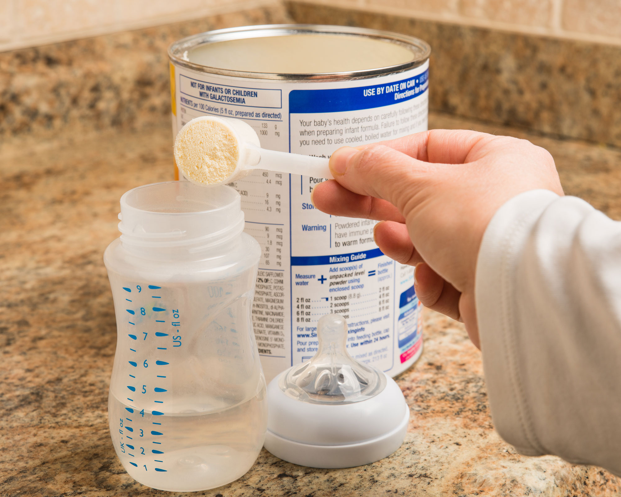 A woman’s hands are holding an empty baby bottle and dumping powdered formula into the bottle