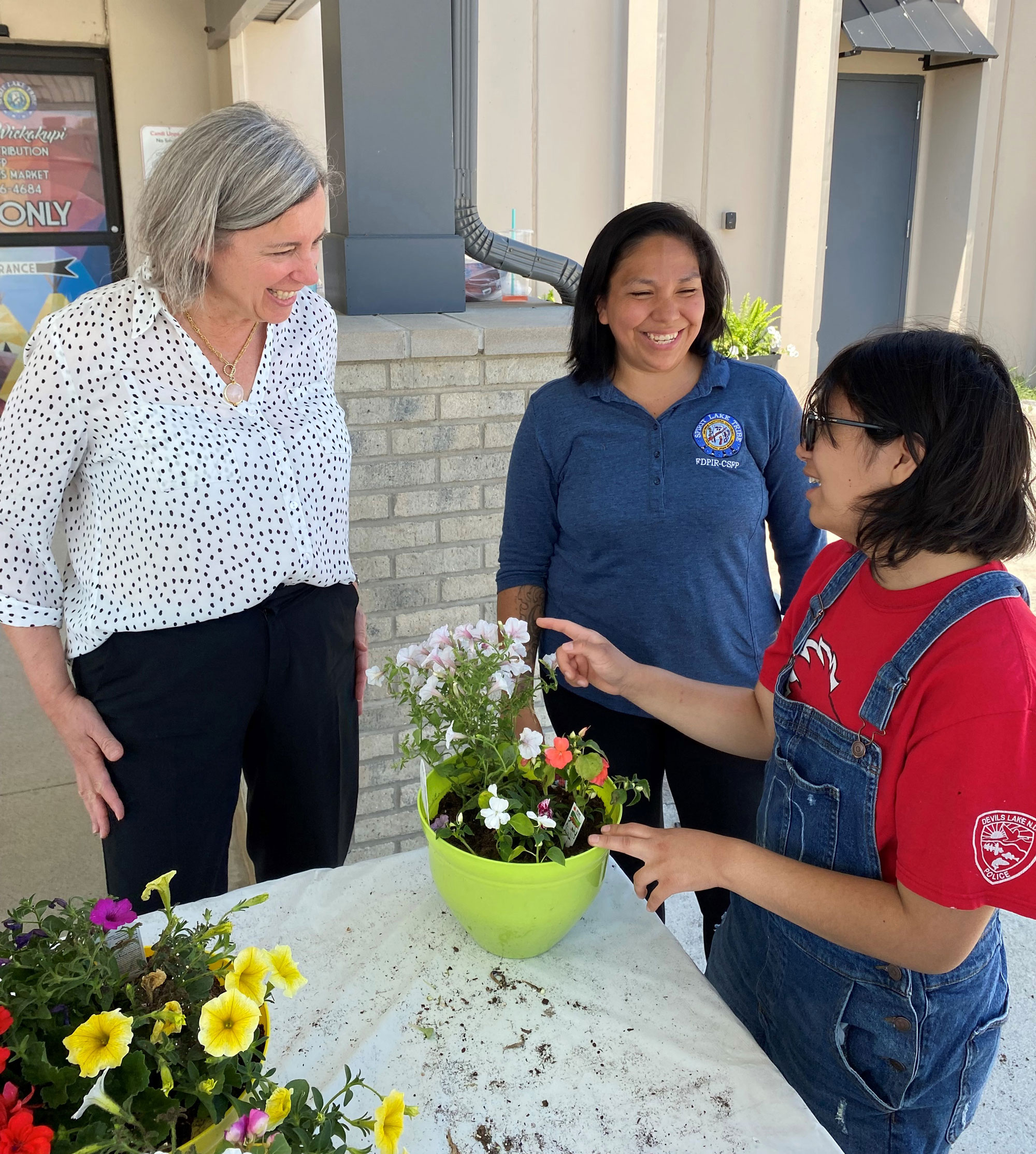 Deputy Under Secretary Stacy Dean smiles with another woman as a child plants flowers in a green pot as part of a youth gardening activity at the Spirit Lake Sioux FDPIR building