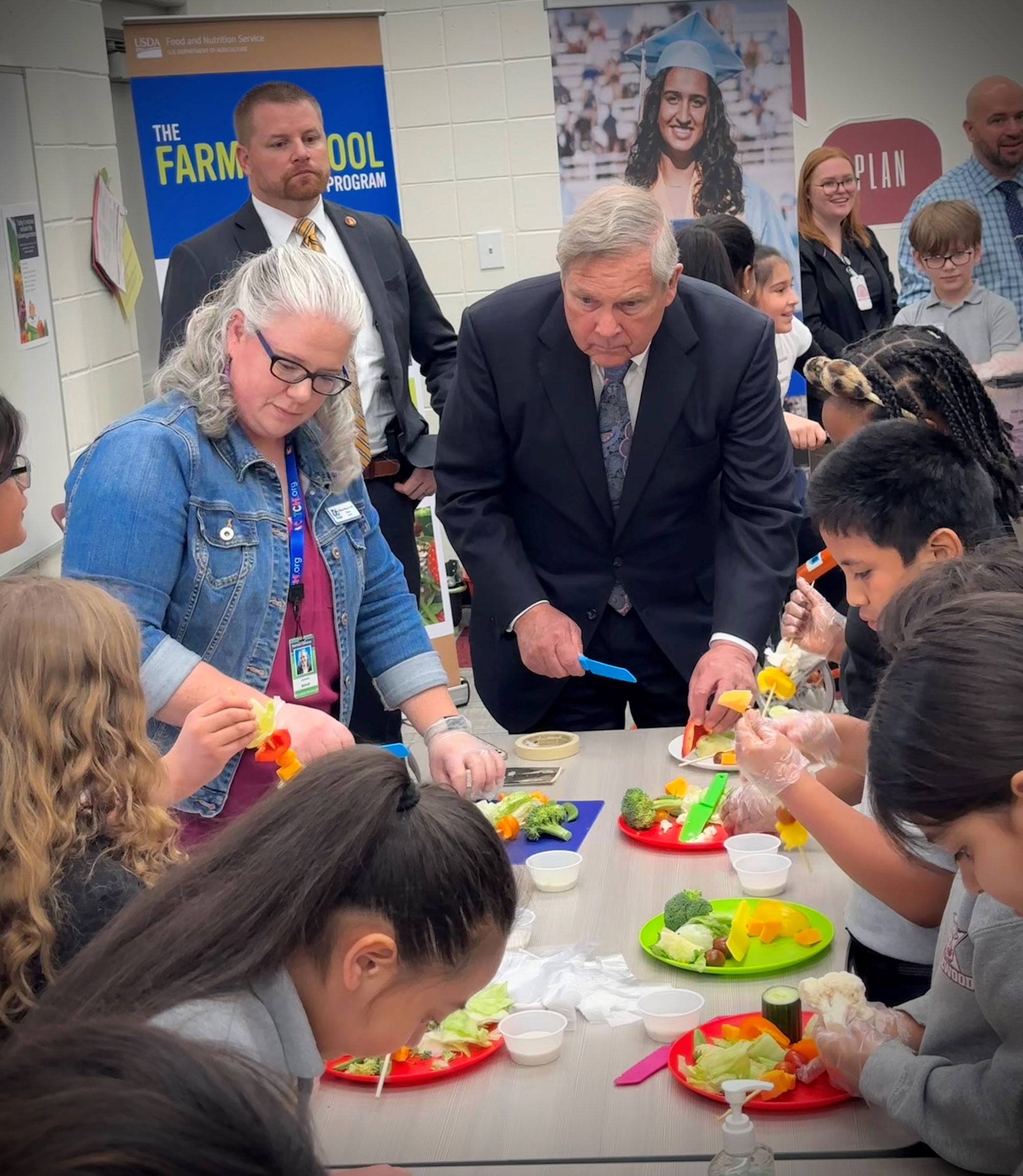 Woman, man and children stand around table and show Secretary Tom Vilsack how to prepare “Salad on a Stick” during a nutrition lesson