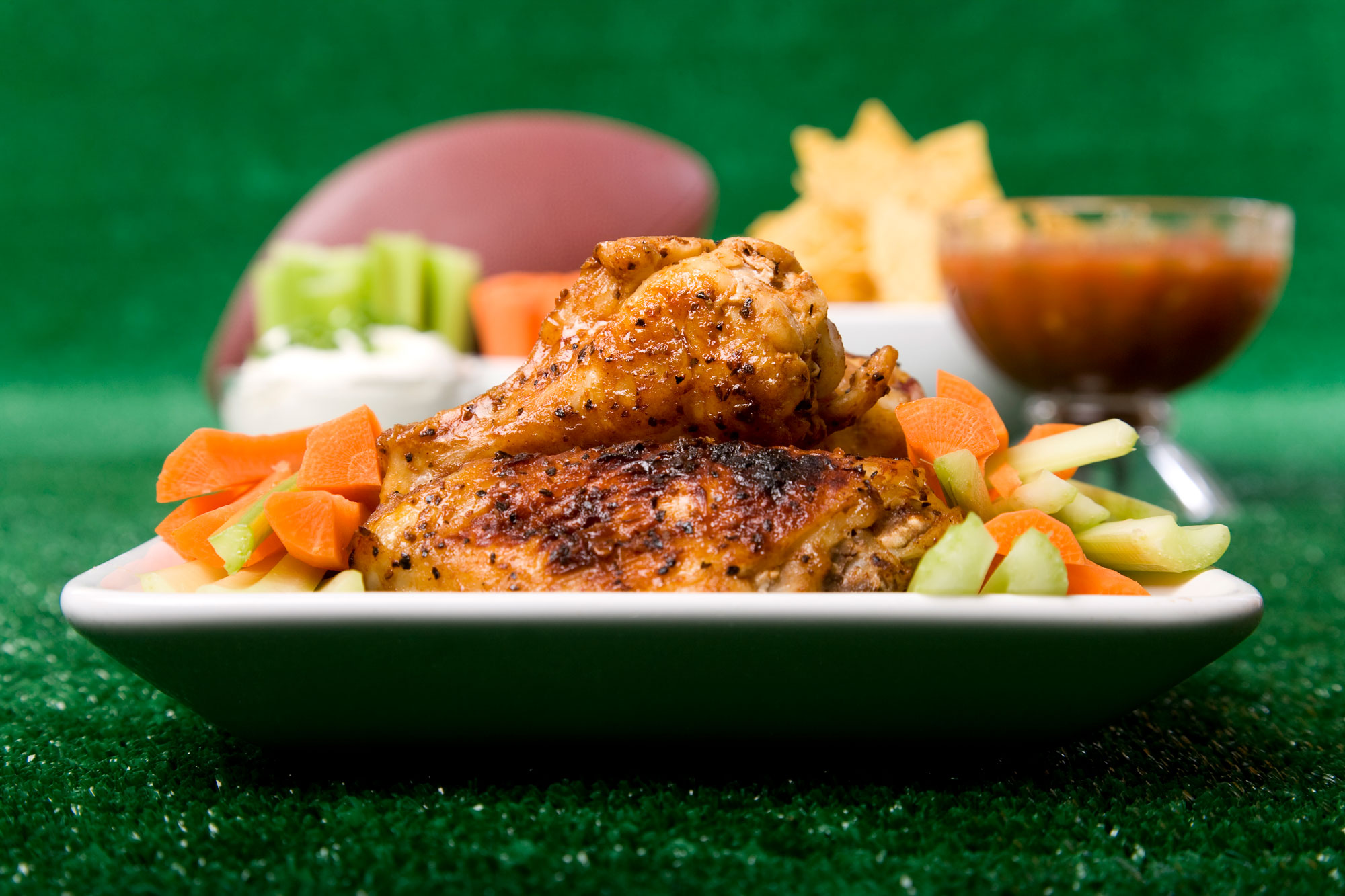 Close up of a full plate of spicy chicken wings with celery and carrot sticks along with championship game party items in the background