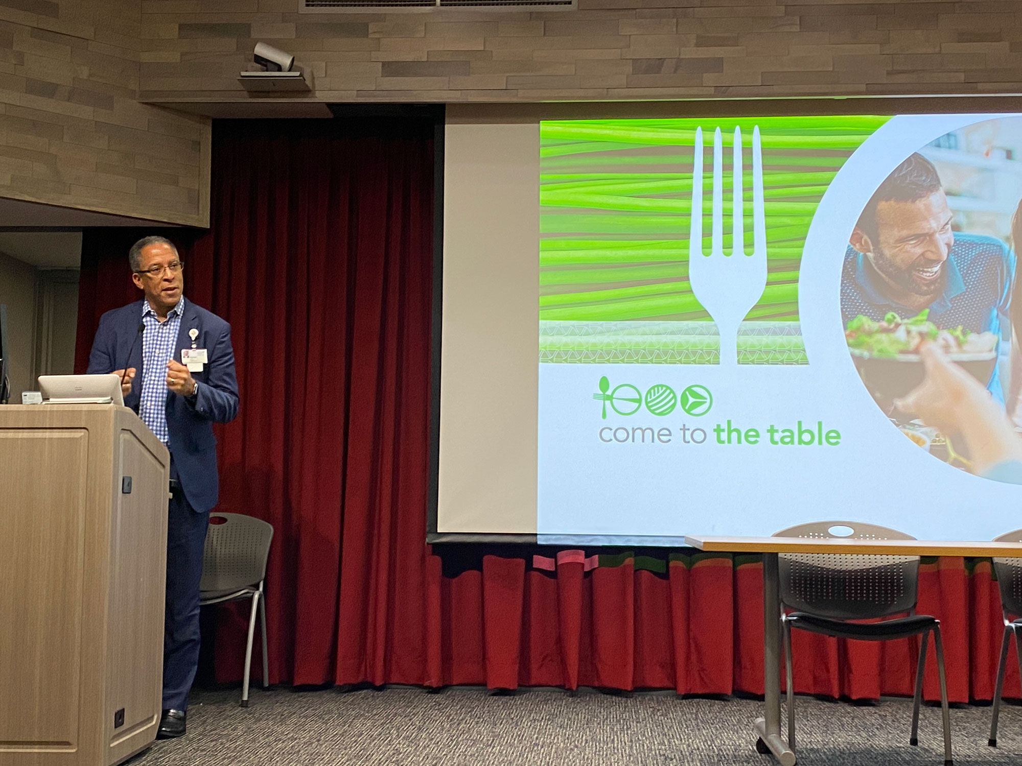 Alonzo Lewis, President of Trinity Health Ann Arbor, standing at a podium giving a presentation. To their left is an image on a screen that reads “come to the table”