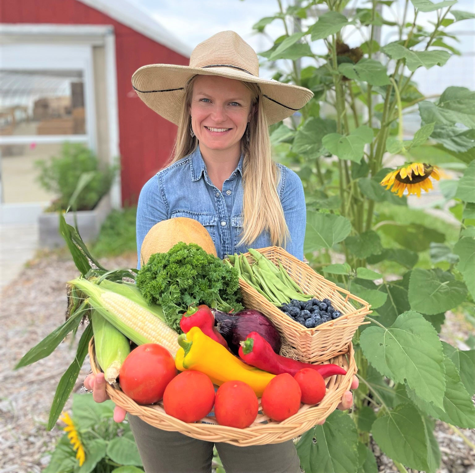 Amanda Sweetman wearing a wide-brimmed straw hat holding a basket of vegetables with sunflowers in the background