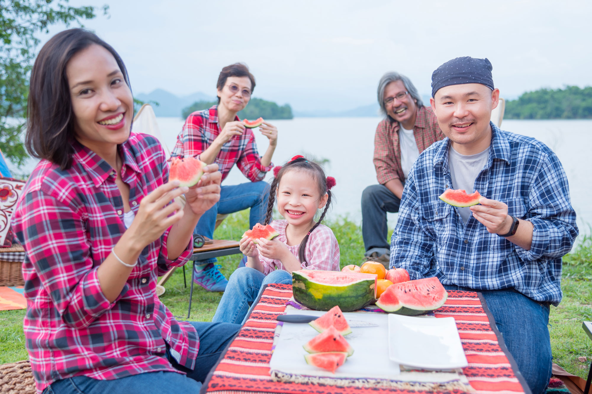 A family eating watermelon outside at a camping site near a lake. A mother, father, and their young daughter are sitting at a table, with two grandparents sitting closer to the lake in the background