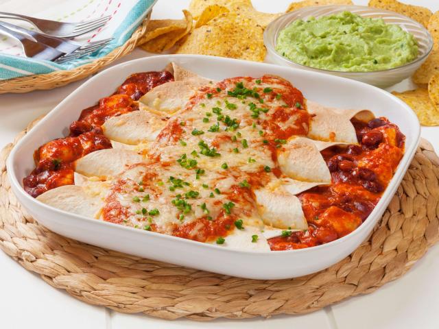 A white serving platter filled with enchiladas rests on a woven trivet. Round tortilla chips, a bowl of guacamole, and utensils are near the platter