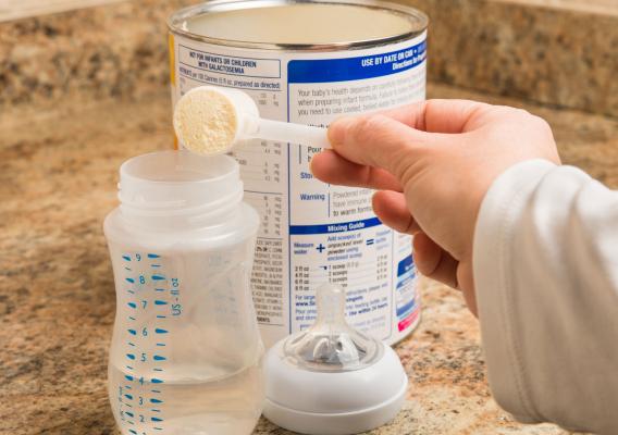 A woman’s hands are holding an empty baby bottle and dumping powdered formula into the bottle