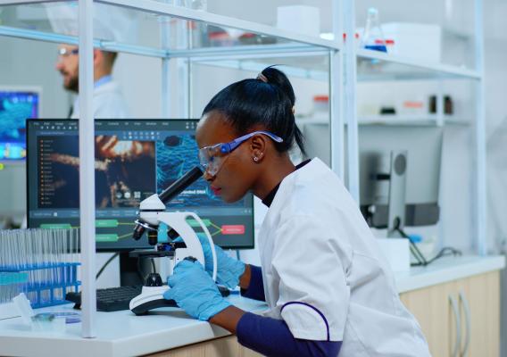 A scientist looks through a microscope in a laboratory. The slide she is observing is magnified on the monitor beside her