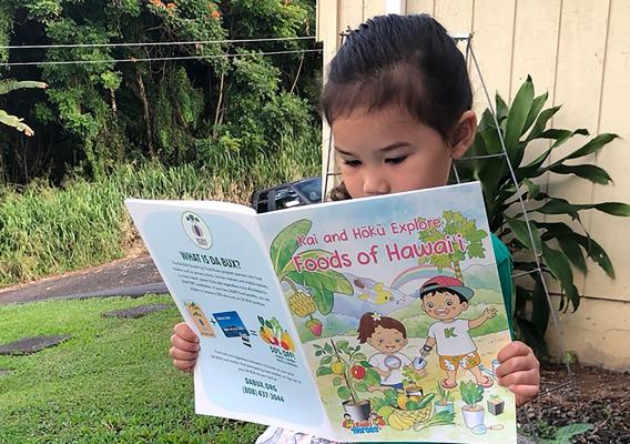 A little girl sitting on a tree stump and reading the book Kai and Hōkū Explore Foods of Hawai‘i