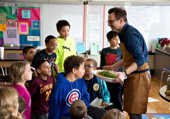 A chef, surrounded by 12 young children in a school classroom, holds a platter of corn, squash, and beans. The children look on with excitement and curiosity