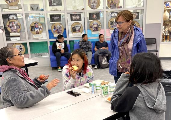Inside a school cafeteria, FNS Administrator Cindy Long is standing in front of a table with two children sitting and eating lunch while another woman is sitting and speaking.
