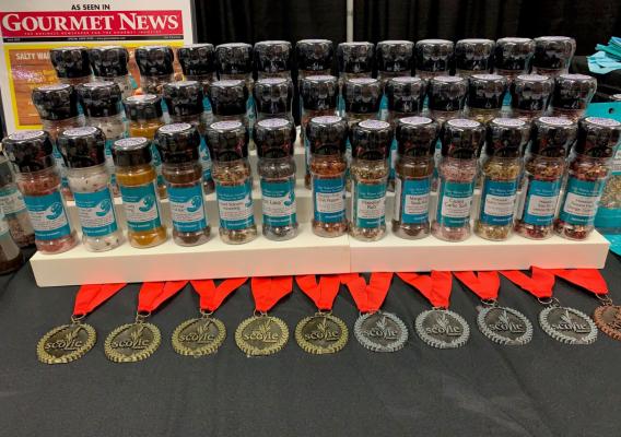 An array of gourmet spices made by Salty Wahine Gourmet Sea Salts are arranged on a three-level stand, with medals placed in front of the stand