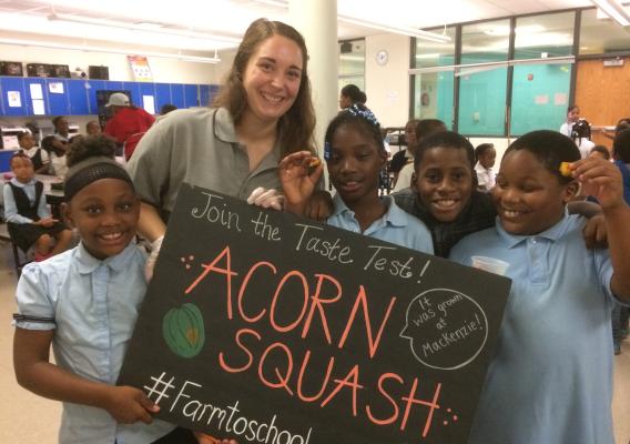 Students holding a sign that says "Join the Taste Test! Acorn Squash. #farmtoschool" and enjoying school-grown produce