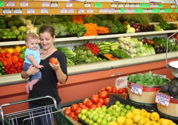 Young mother with baby selects items in produce aisle of grocery store.