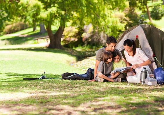 Family of four reading on the grass in front of a tent with tree in background. Photo courtesy of ThinkStock.