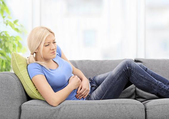 Blond woman with a painful expression sitting on a grey sofa at home with her hands placed on her stomach