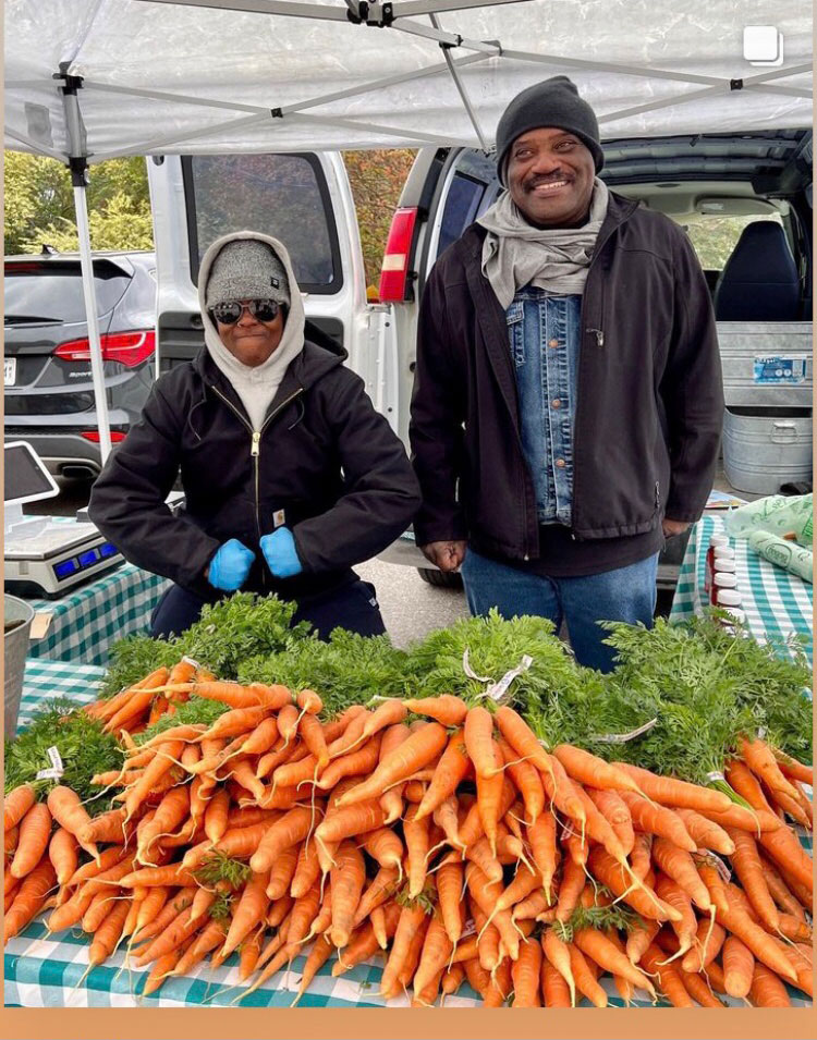 A man smiling beside a woman posing her arms in front of a table with carrots piled up